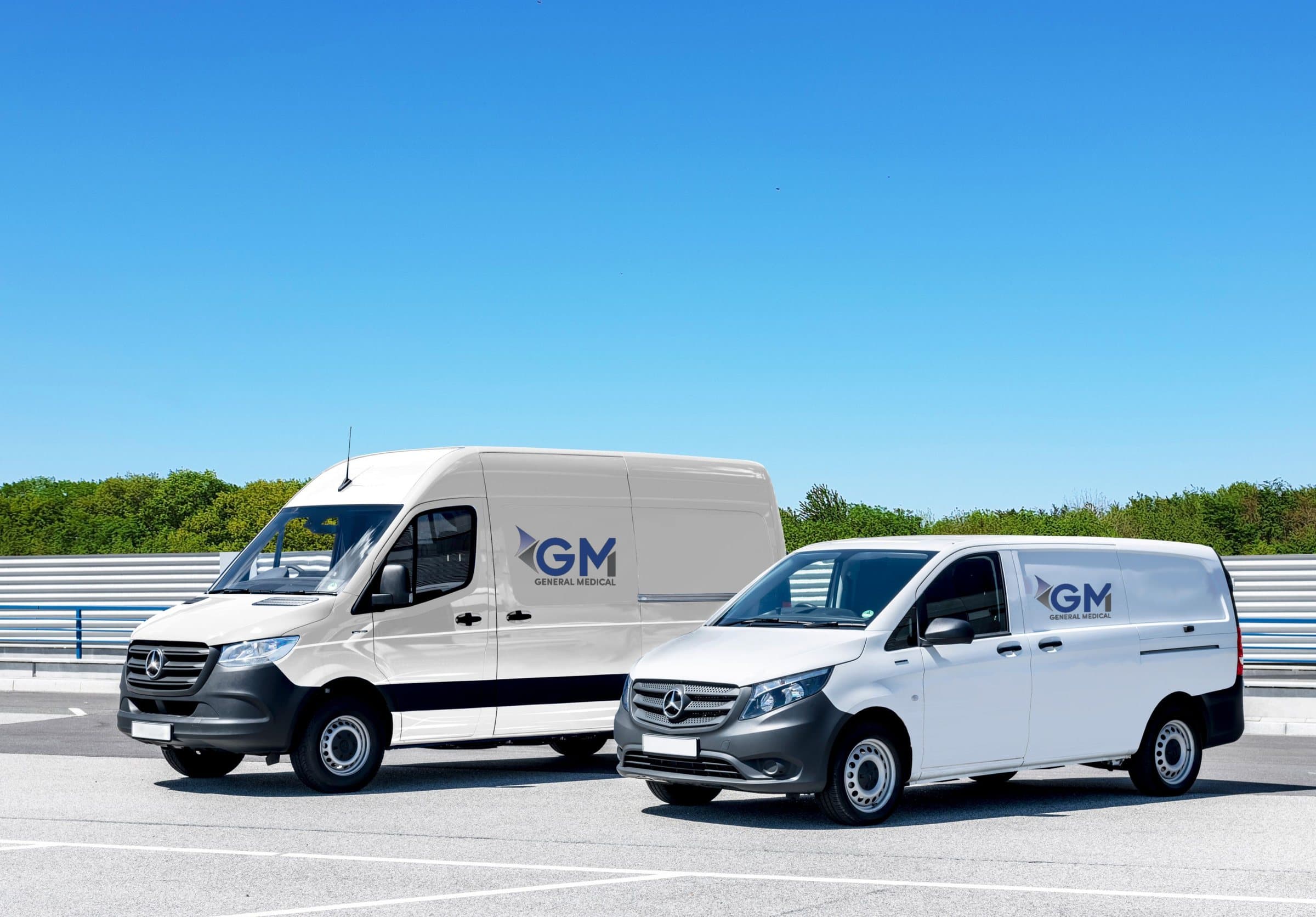 Two white General Medical transport vans, each with the GM logo on its sliding door, parked side by side under a clear blue sky.