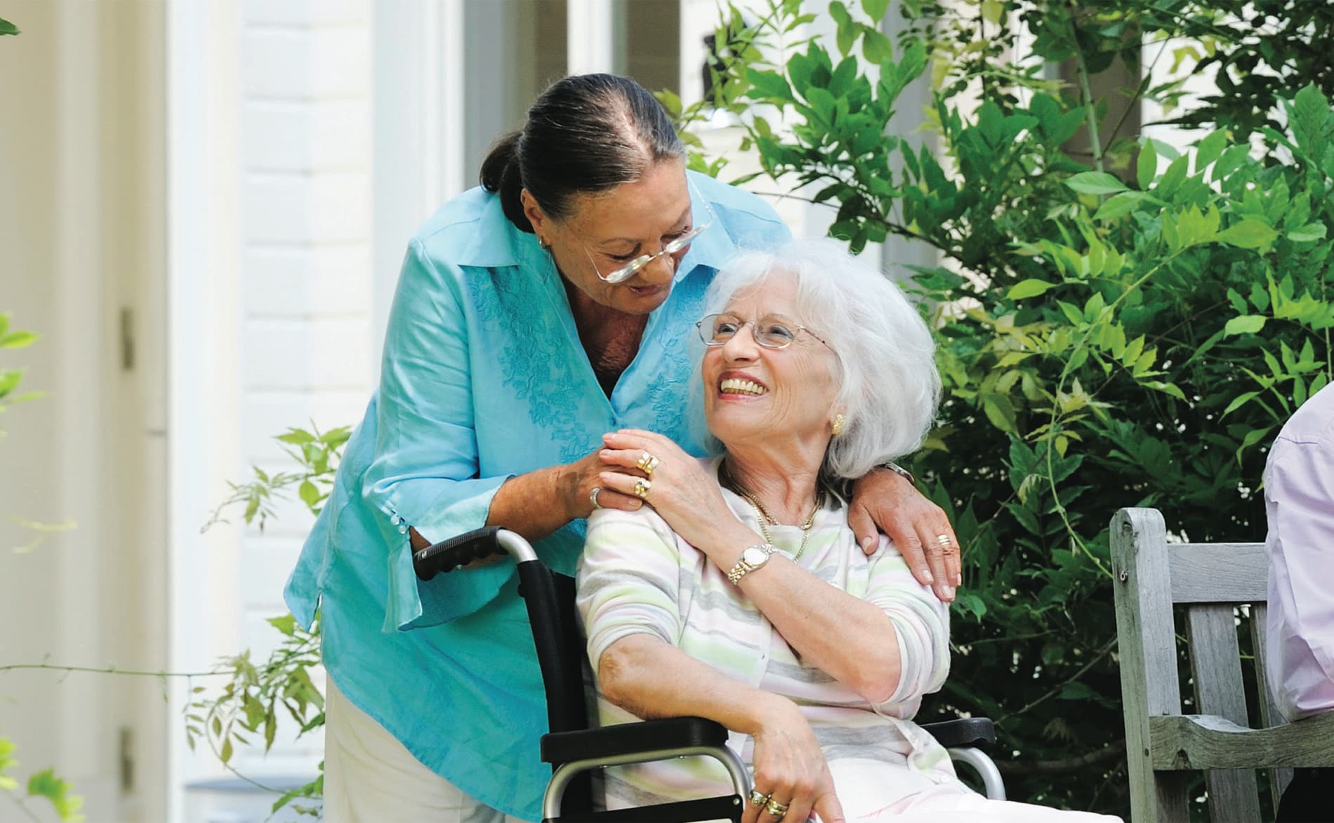 A caregiver leaning in to hug a smiling older woman seated in a wheelchair on a leafy porch.