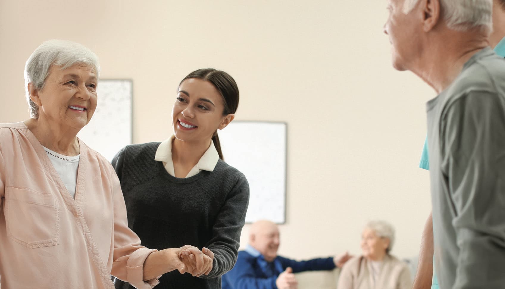 A smiling caregiver holding hands with an older woman in a bright common room while a colleague supports another resident.