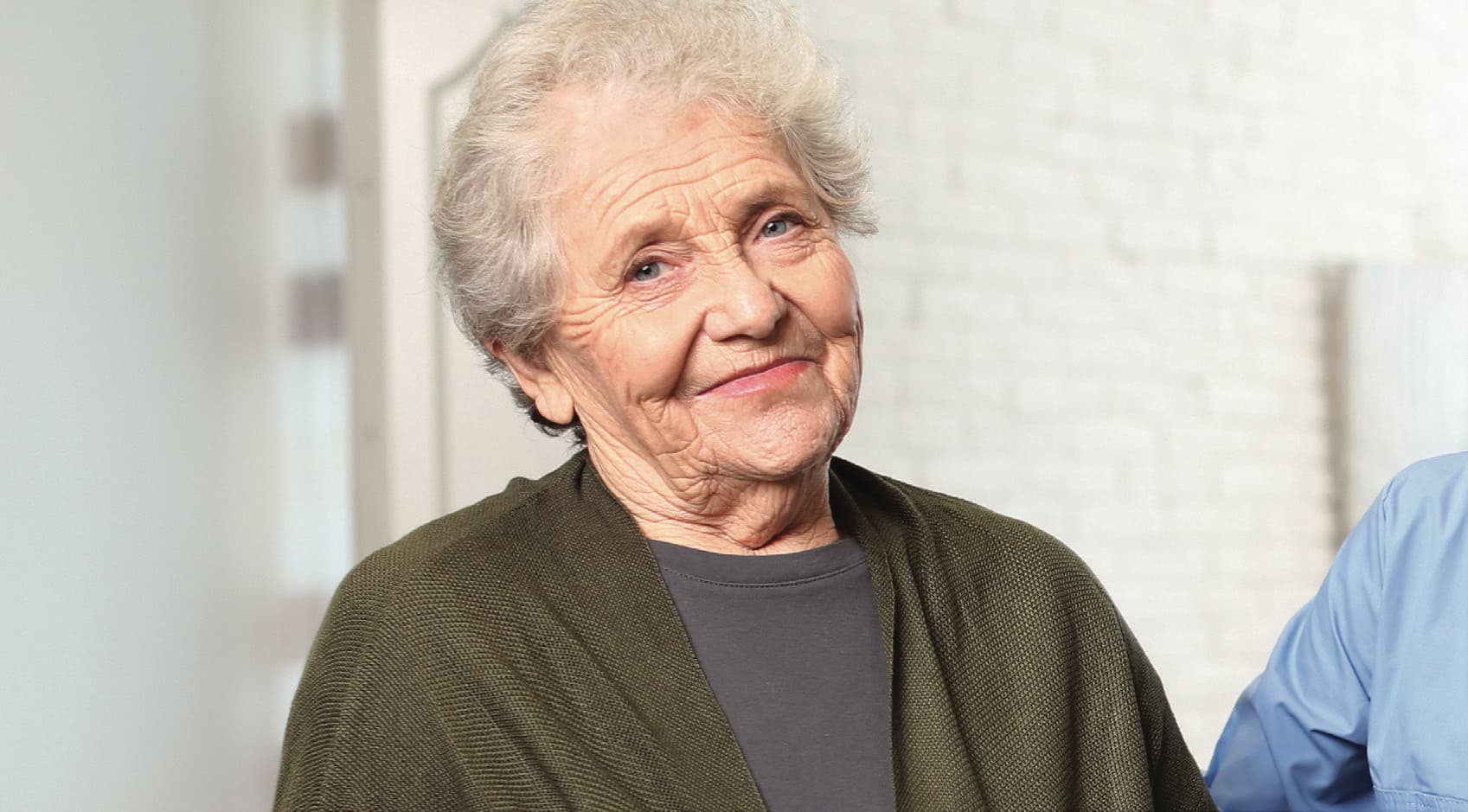 An older woman in a green cardigan smiling warmly, with a clinician in blue scrubs beside her.